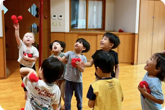 木目調の保育室(遊戯室)で、上を見上げながら一斉に赤いお手玉を投げて遊ぶ多くの子どもたちの様子