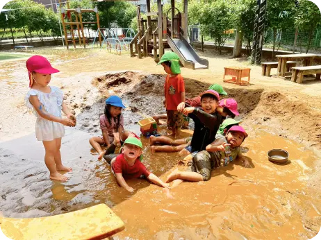 晴れた日の園庭で、全身泥だらけになりながら泥水の中で元気に遊ぶ多くの子どもたちの様子