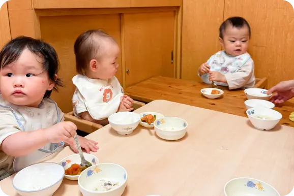 木目調の保育室で、エプロンをつけて幼児用椅子に座り、給食や離乳食を食べる乳幼児たちの様子