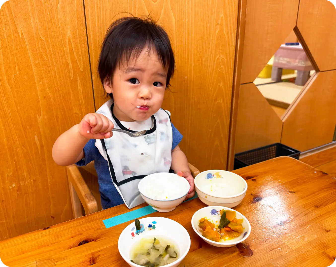 子ども用の椅子に座り、スタイをつけて自分でスプーンを持って給食を食べる男の子の様子