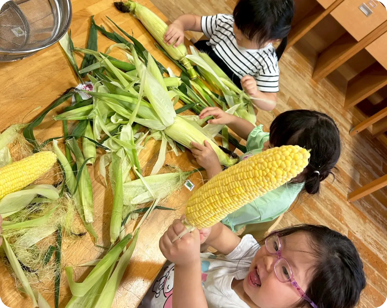 木製テーブルの上いっぱいに置かれたとうもろこしの皮を、みんなで一緒にむいて楽しむ子どもたちの様子