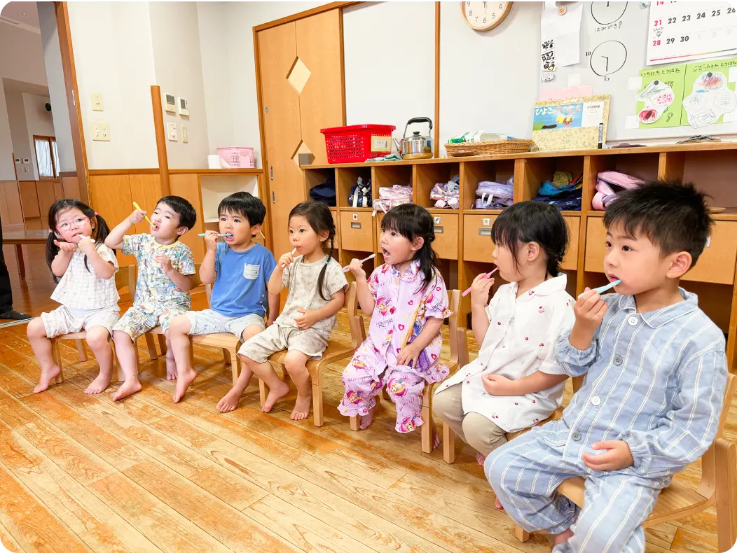 木目調の保育室で、保育士の後ろに並んで一列になり、体を動かして室内遊びを楽しむ子どもたちの様子