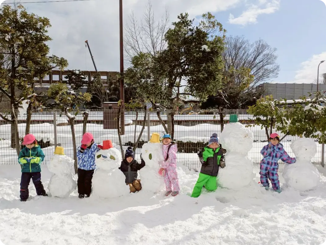 雪が積もった屋外で、防寒着を着て大きな雪だるまをいくつも作り、その横に並んで立つ子どもたちの様子