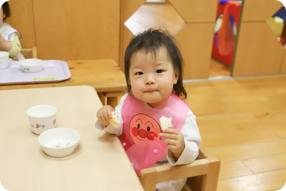 木目調の保育室で、エプロンをつけて幼児用椅子に座り、パンを食べておやつ(食事)を楽しむ乳児の様子